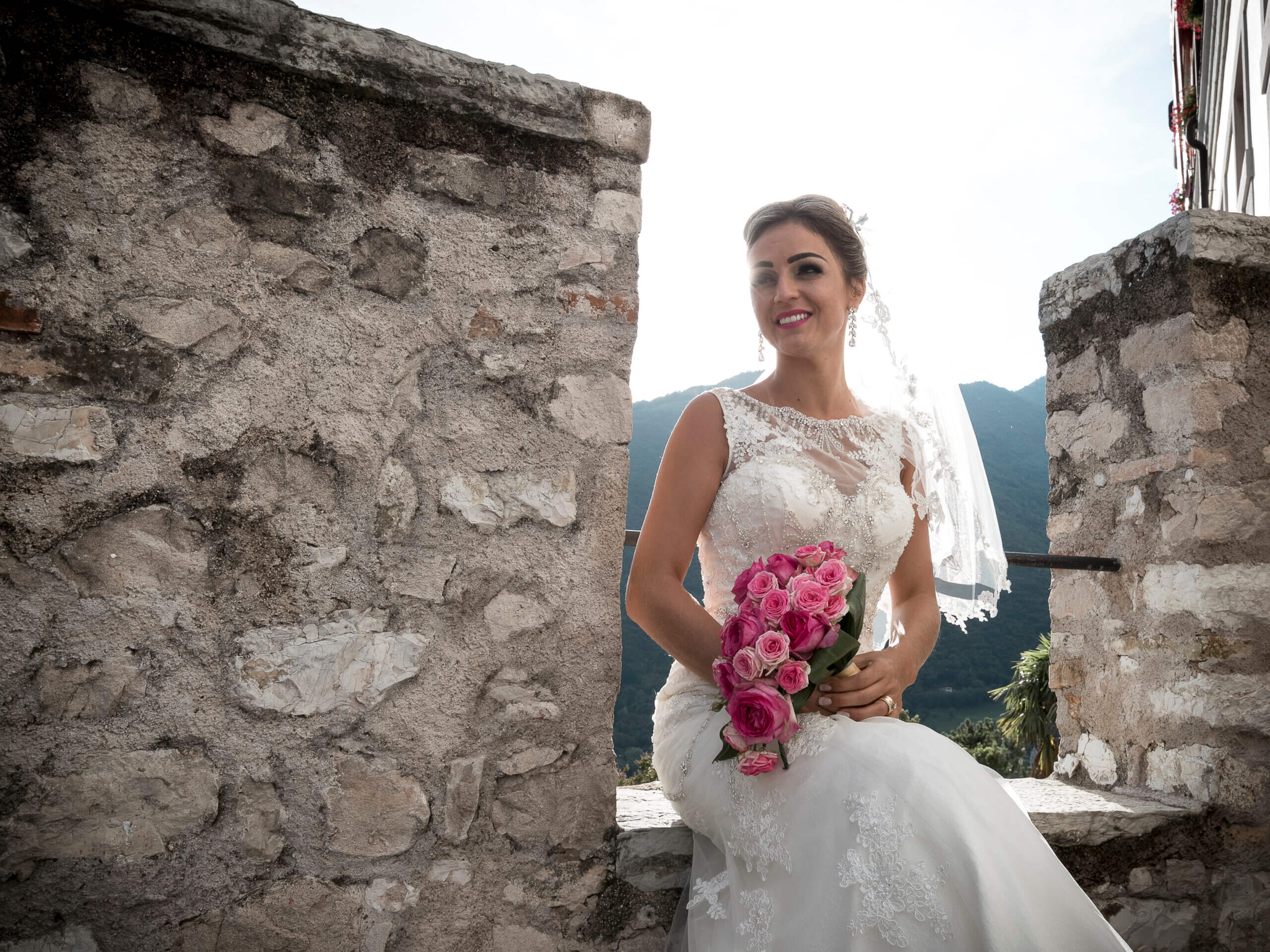 Lächelnde Braut die auf der Mauer sitzt mit einem wunderschönen Brautstrauss in der Hand.