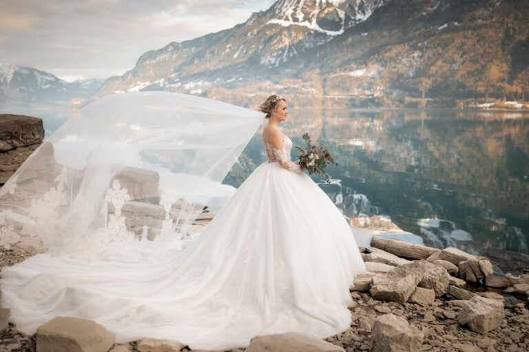 Brautkleid Shooting von Monova Bridal am Wasser in den Bergen. Der Schleier weht im Wind.